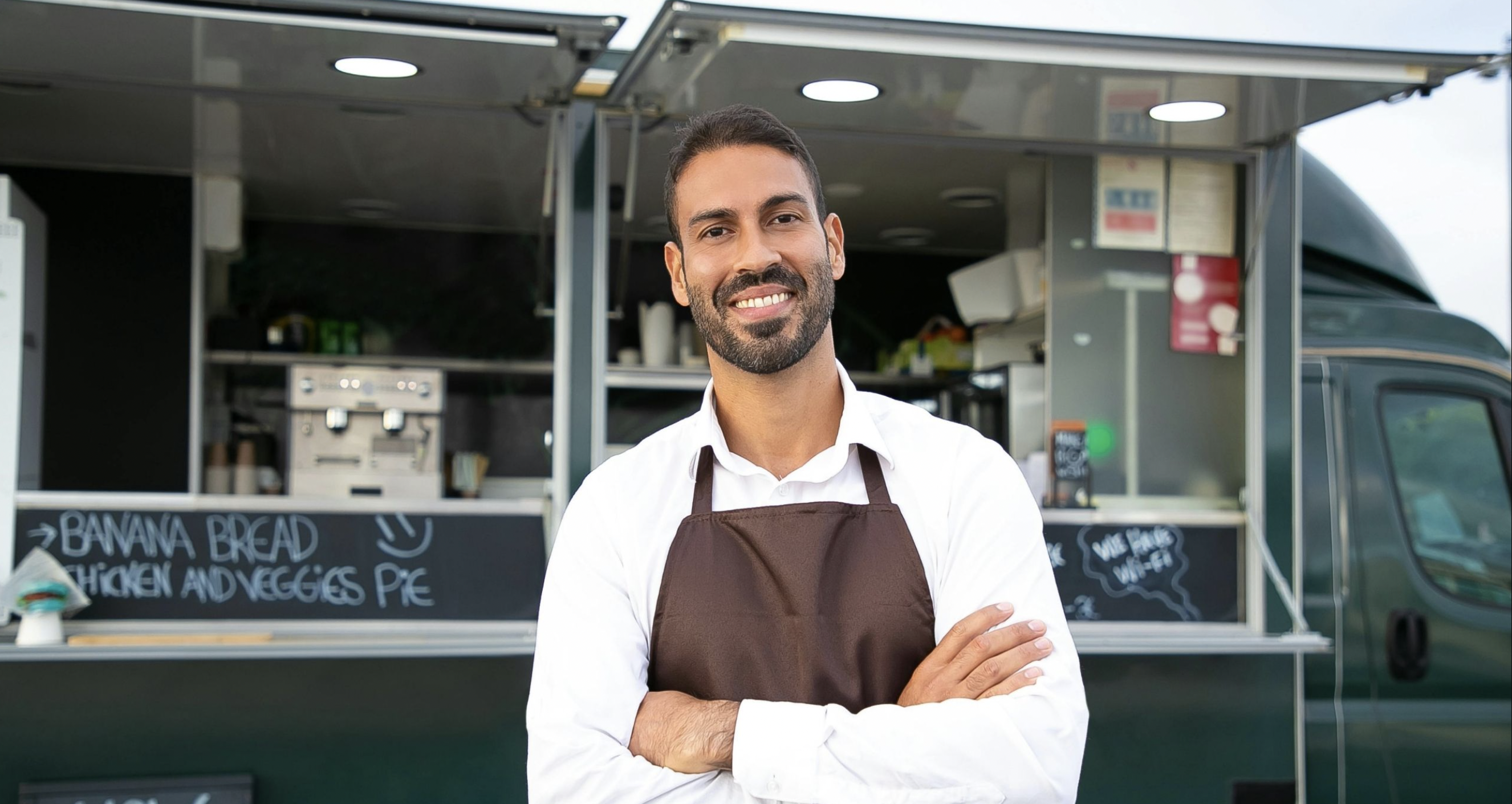 Smiling food truck owner in apron standing with arms crossed in front of his mobile kitchen