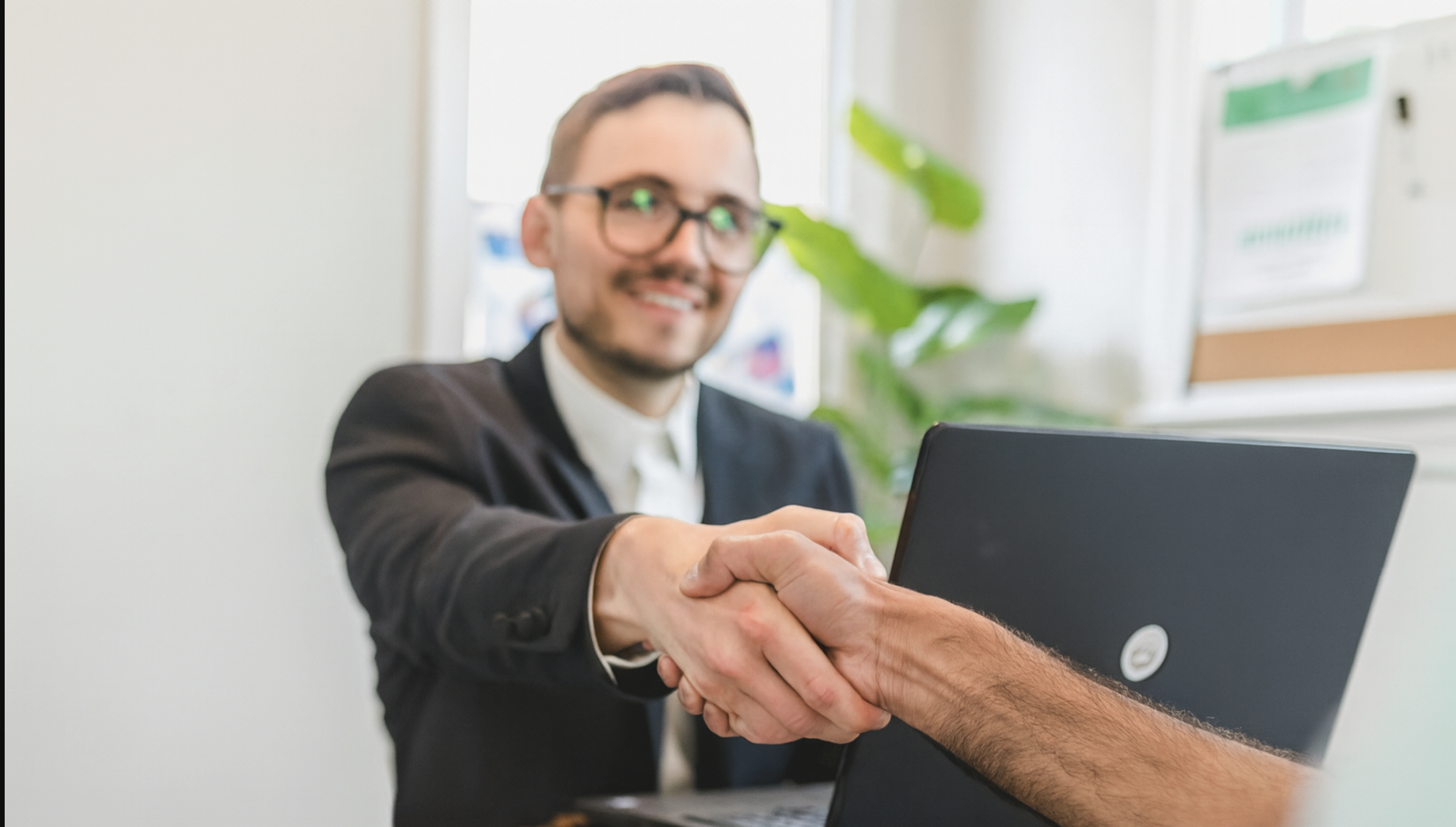 A professional man in a suit shaking hands with a client across a desk with a laptop on it, in a bright office setting.