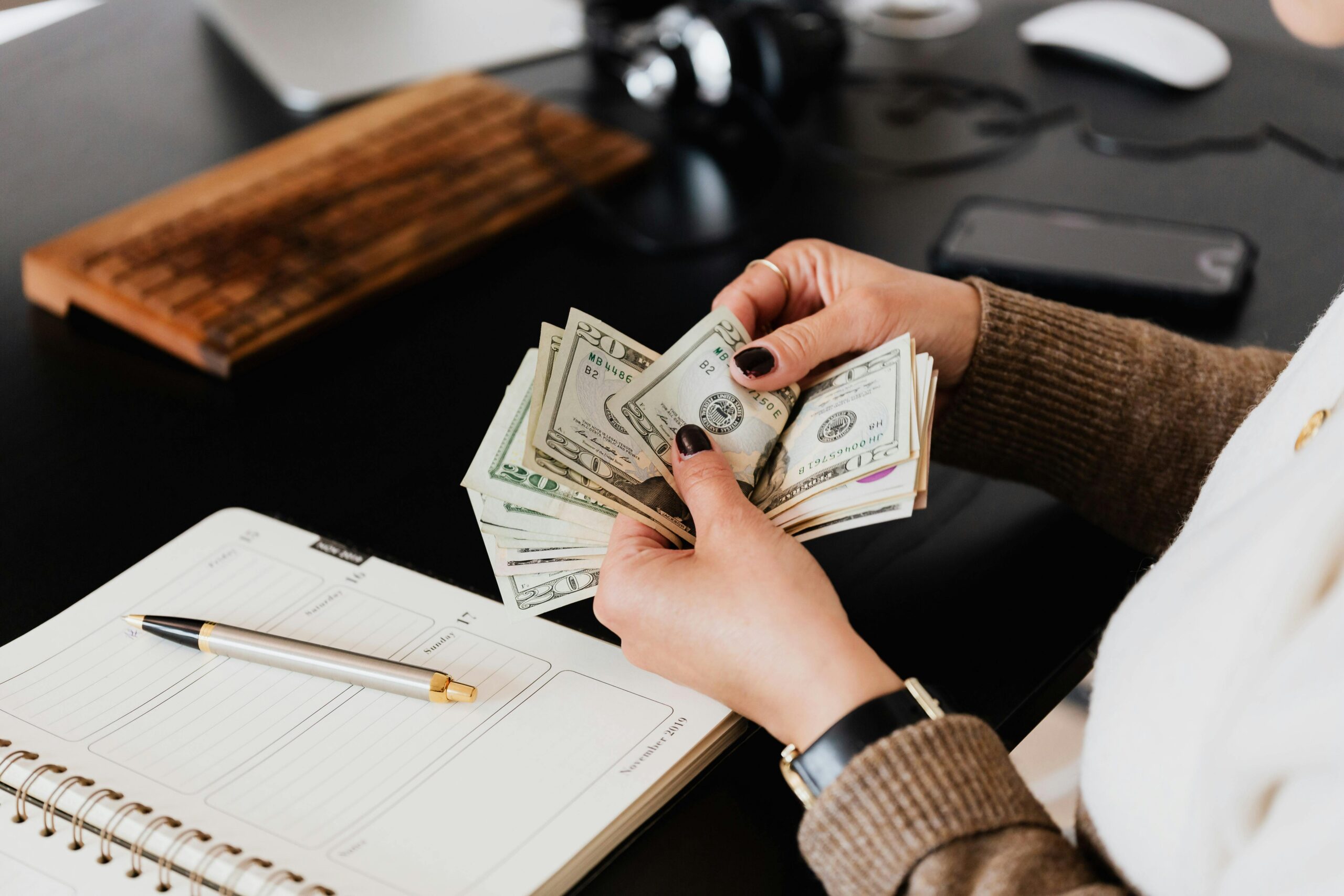 Person counting U.S. dollar bills at a desk with a notebook, pen, and computer keyboard in view.