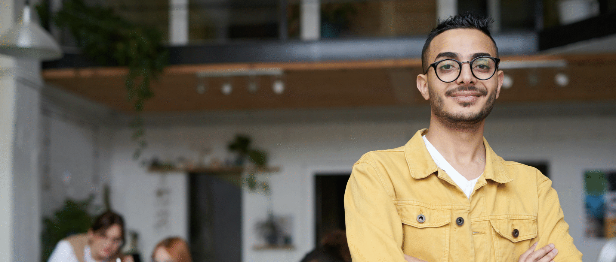 Confident young professional standing with folded arms in a modern office while colleagues collaborate at a desk in the background.