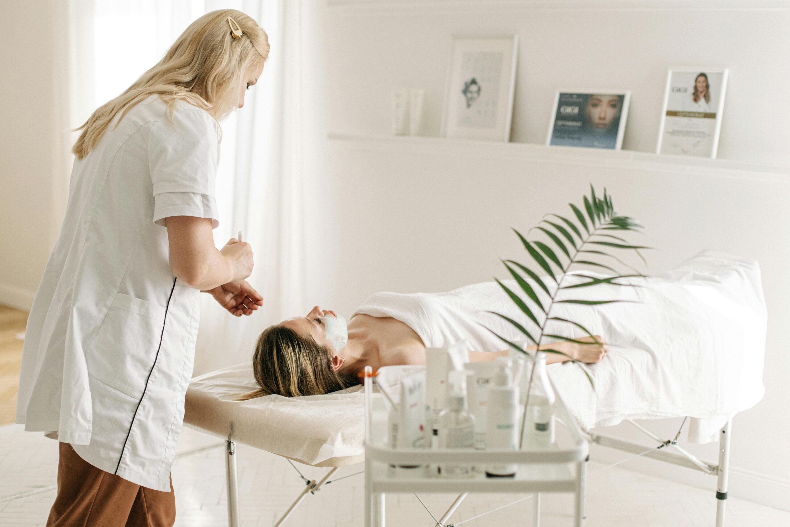 Skincare professional applying a facial treatment to a client lying on a spa bed in a bright, minimalist treatment room.