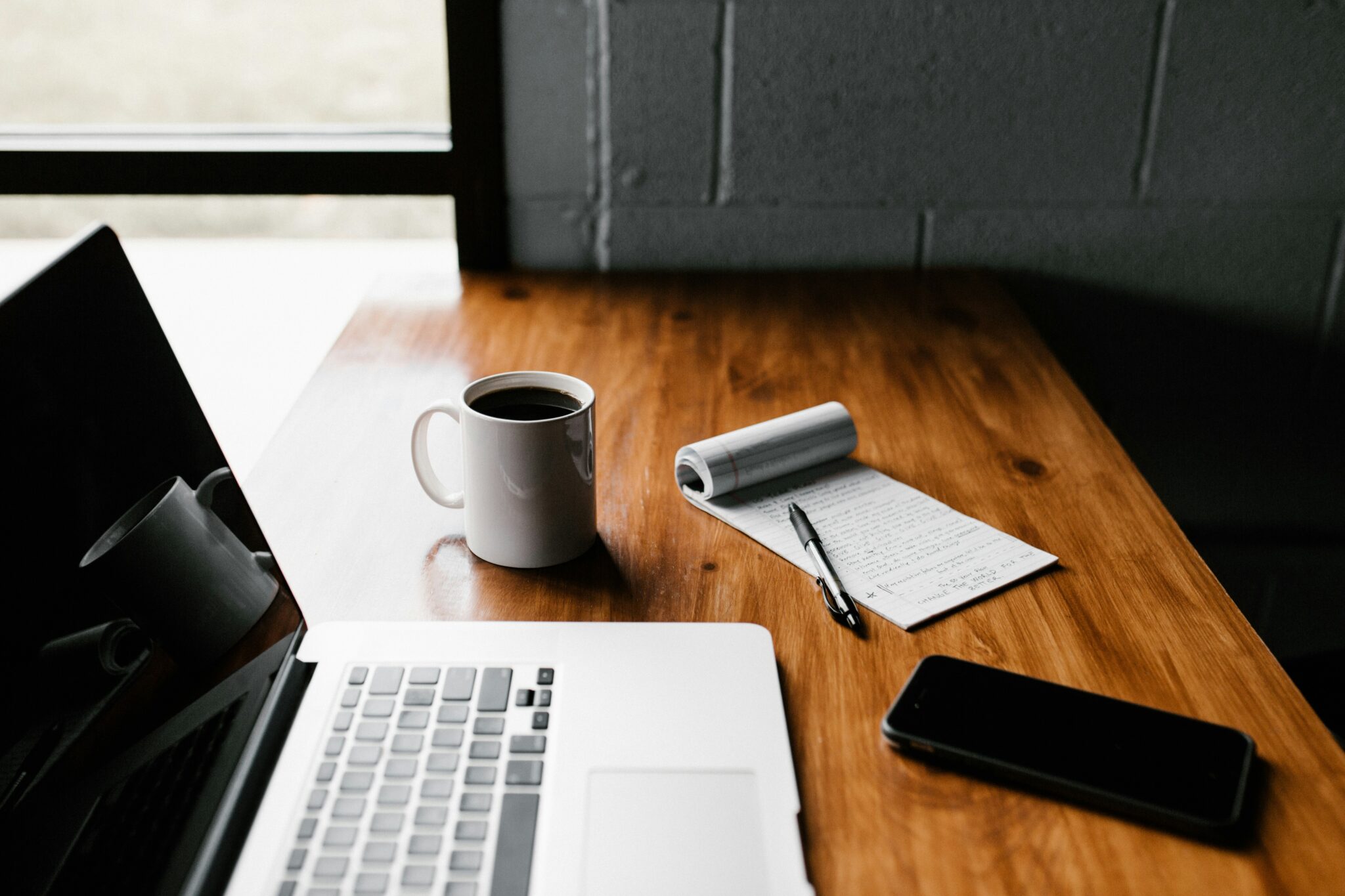Wooden desk setup with an open laptop, a cup of coffee, a notepad filled with handwritten notes, a black pen, and a smartphone near a window.