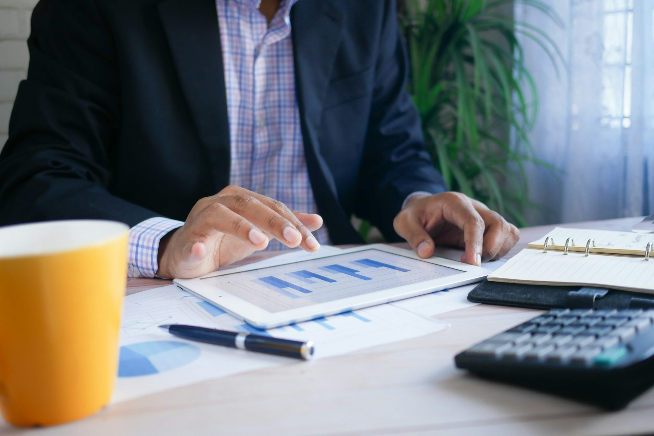 Business professional analyzing financial charts on a tablet at a desk, surrounded by documents, a calculator, a notebook, and a yellow coffee mug.